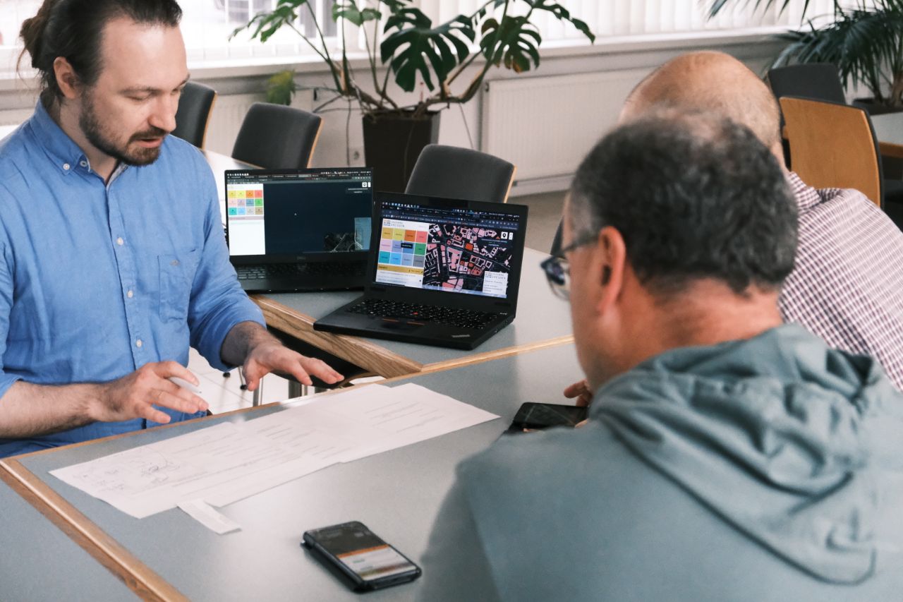 Man sieht Menschen beim Usabilty-Test zusammensitzen. Sie blicken gemeinsam auf Abbildungen, die auf dem Tisch liegen. Im Hintergrund stehen Laptops auf einem Tisch, man sieht Stühle und Tische und Pflanzen vor einer Fensterfront.