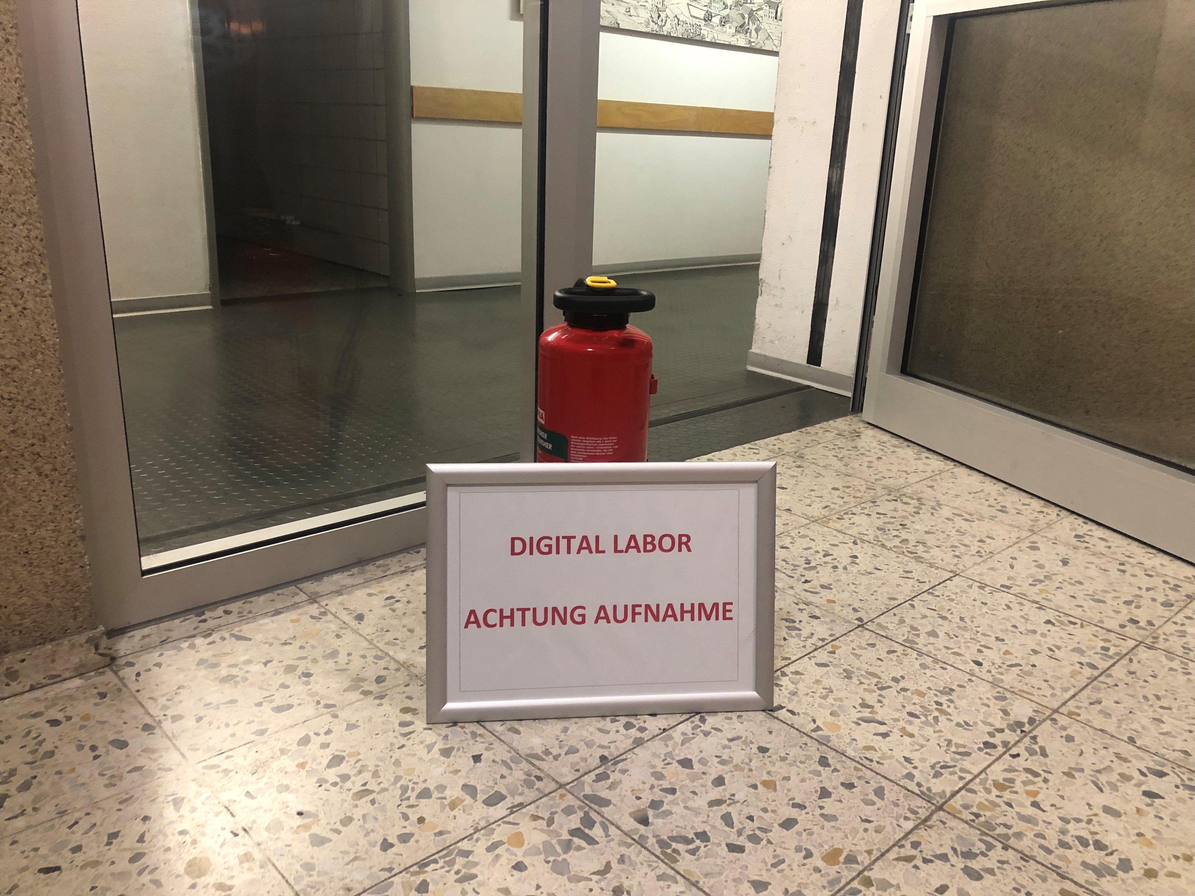 Auf dem Bild steht ein Schild mit der Aufschrift "Digital Labor - Achtung Aufnahme" an einen Feuerlöscher gelehnt vor einer Glastür.