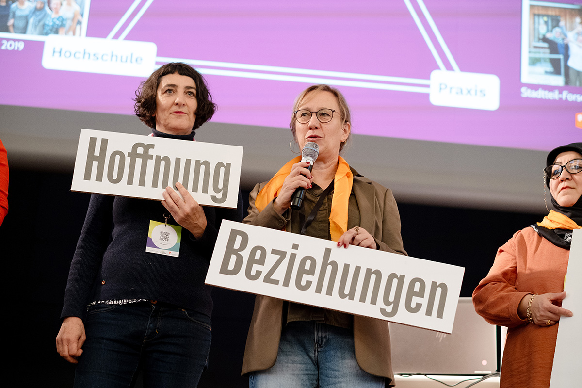 Zwei weiblich gelesene Personen stehen vor einer Präsentation. Die rechte Person spricht in ein Mikro und hält ein Pappschild mit der Aufschrift Beziehungen in der Hand. Die Linke Person hält ein Schild mit der Aufschrift Hoffnung in der Hand.